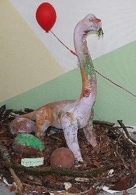 Langhalsiger Modell-Dinosaurier in Nest aus Zweigen mit Eiern und rotem Luftballon