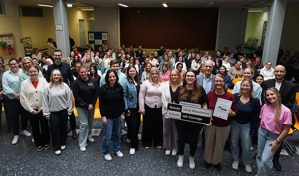 Große Gruppe von Schüler*innen in einer Aula, viele lächelnd; vorne halten mehrere Personen ein Schild „Schule ohne Rassismus — Schule mit Courage“.